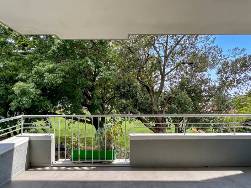 A spacious balcony featuring tiled flooring and a metal balustrade, offering a pleasant view of mature trees and greenery.