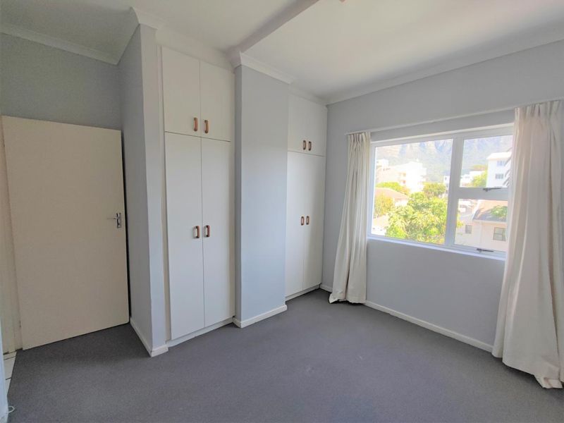 A bright, empty bedroom featuring neutral walls, carpeted flooring, and ample built-in cupboard storage with a view through the window.
