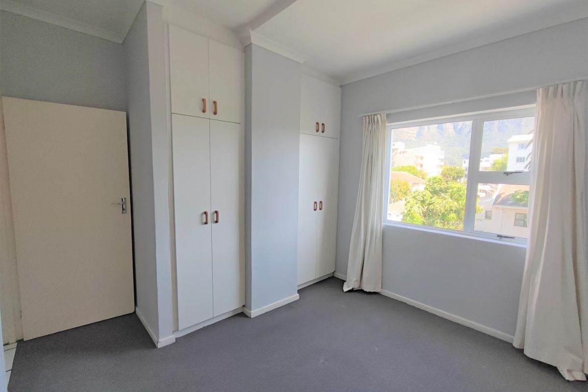 A bright, empty bedroom featuring neutral walls, carpeted flooring, and ample built-in cupboard storage with a view through the window.