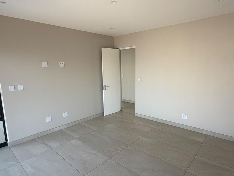 A bright, empty bedroom featuring neutral wall tones, large format floor tiles, recessed lighting, and a modern black-framed glass door.