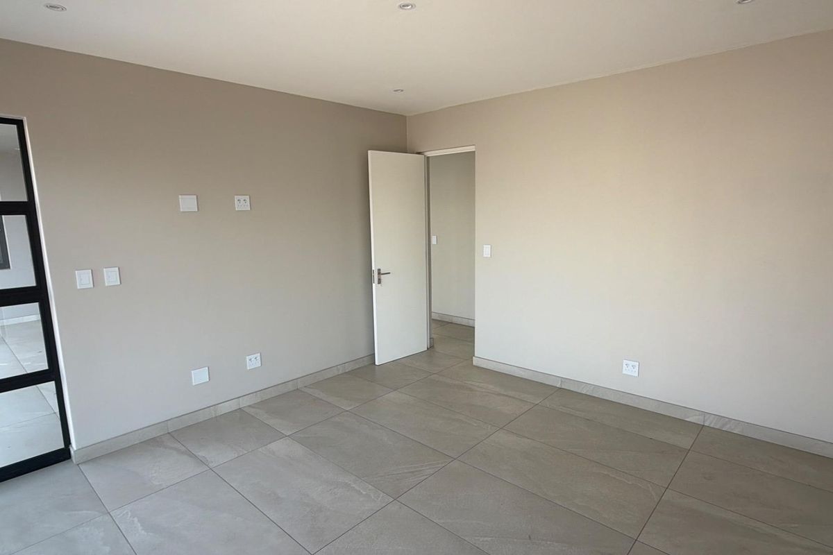 A bright, empty bedroom featuring neutral wall tones, large format floor tiles, recessed lighting, and a modern black-framed glass door.