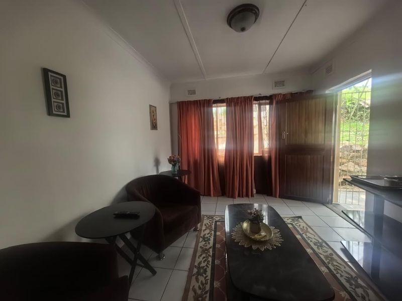 A living room featuring tiled flooring, a ceiling light fixture, and direct access to the exterior through a timber door with a security gate.