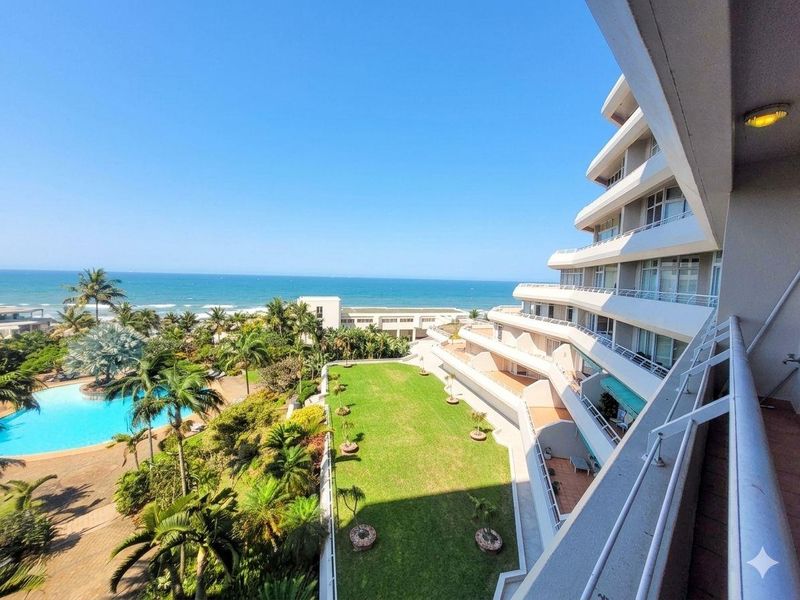 Elevated view from a balcony overlooking the apartment complex grounds, swimming pool, landscaped gardens, and the ocean.