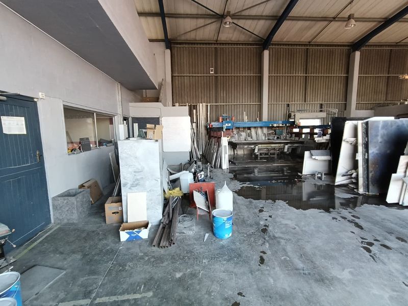 Industrial warehouse space featuring high ceilings with exposed steel trusses, concrete flooring, and an internal office window, currently used for stone fabrication.