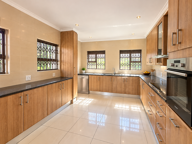 A well-maintained kitchen featuring wood-finish cabinetry, granite countertops, integrated stainless steel appliances, tiled flooring, and ample natural light through security-barred windows.