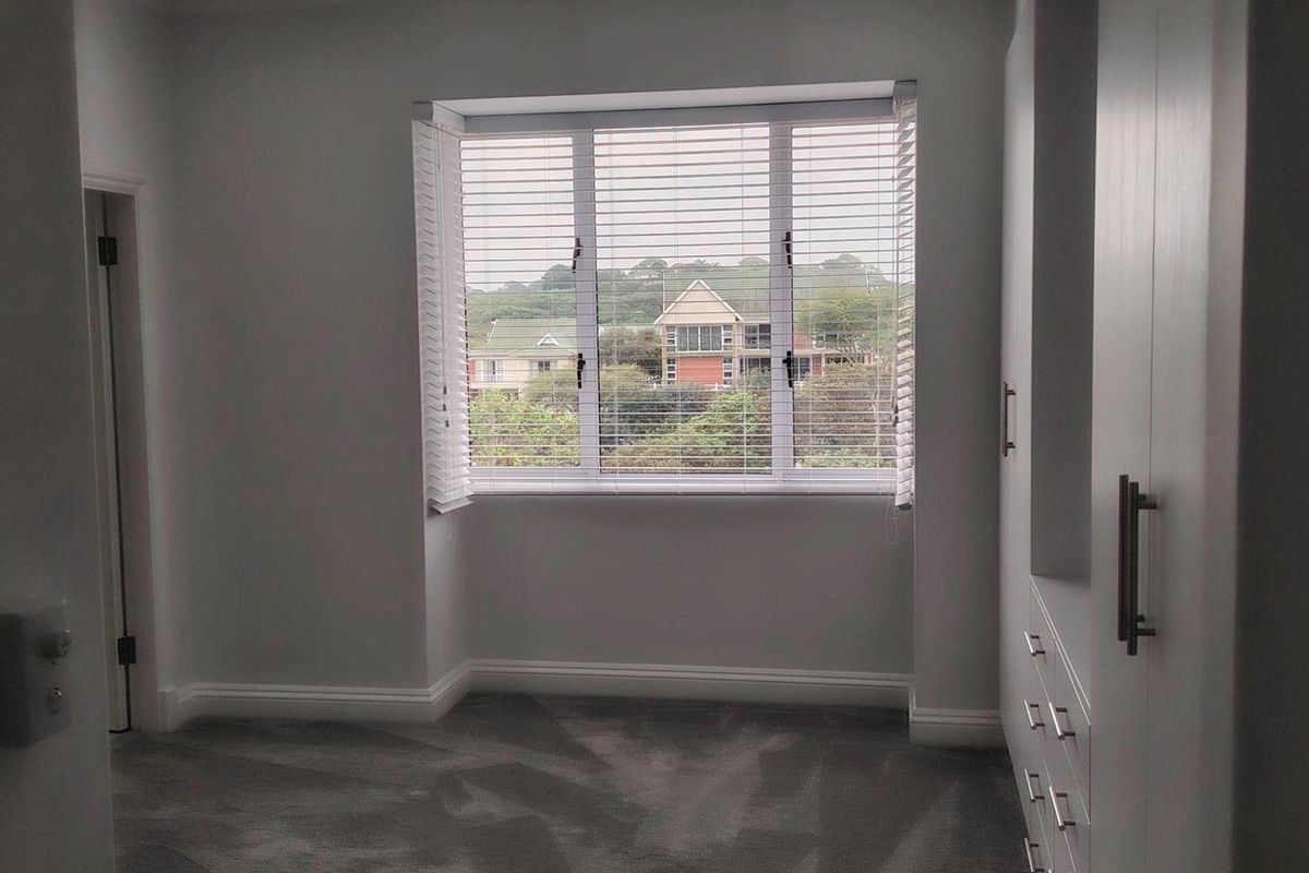 A bright, empty bedroom featuring neutral carpeting, built-in storage cupboards, and a large window with venetian blinds offering a pleasant neighbourhood view.