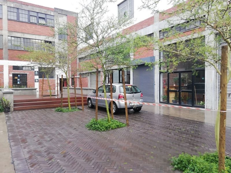 A modern industrial-style courtyard featuring brick paving, timber decking, and exposed brick architecture with large steel-framed windows and roller shutter access.