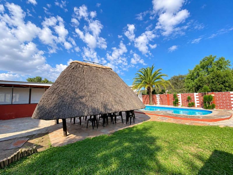 Outdoor entertainment area featuring a traditional thatch lapa, swimming pool, and stone-paved patio set within a landscaped garden with brick boundary walls.