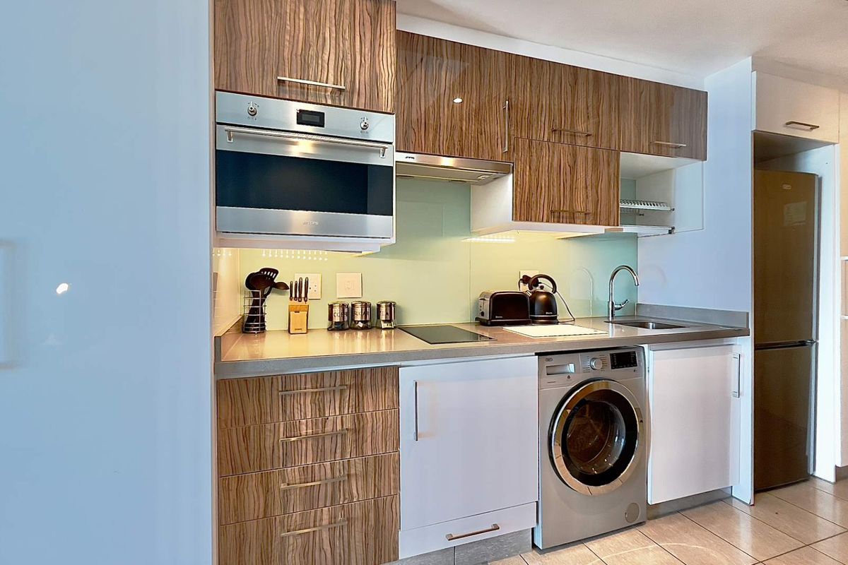 Compact modern kitchen featuring wood-grain cabinetry, integrated oven, glass cooktop, stainless steel sink, and a built-in washing machine.