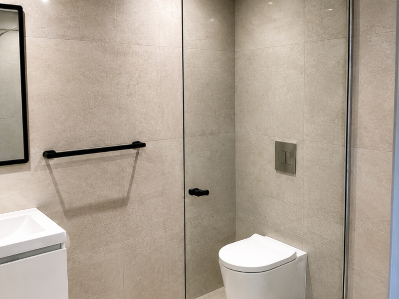 Modern bathroom featuring a wall-mounted toilet, glass shower screen, vanity basin, and neutral floor-to-ceiling tiling with contemporary black fixtures.