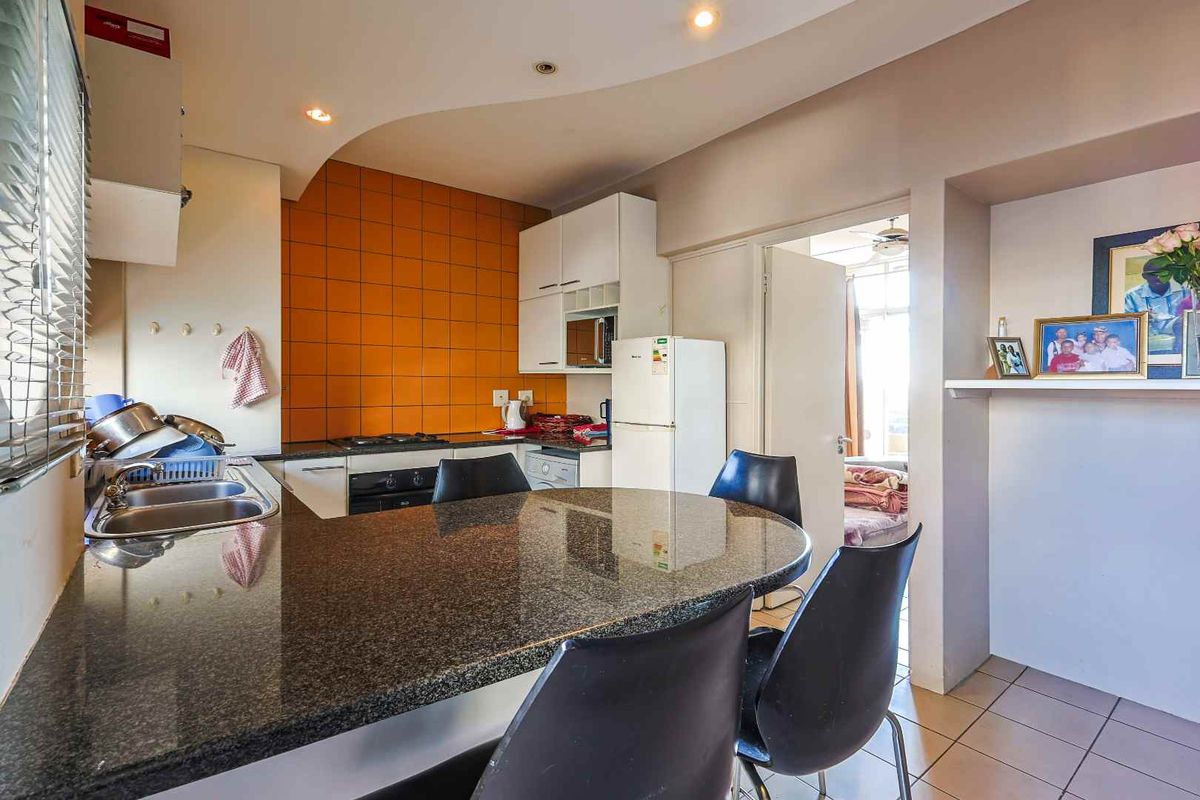 Compact kitchen featuring a granite breakfast bar, built-in oven and hob, and orange tiled splashback, opening into an adjacent living area.