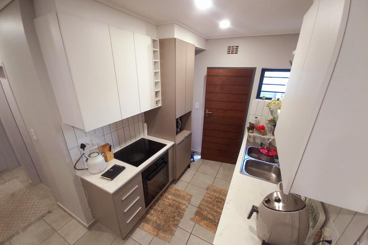 Modern galley-style kitchen featuring white and taupe cabinetry, integrated electric hob, built-in oven, double stainless steel sink, and tiled flooring.