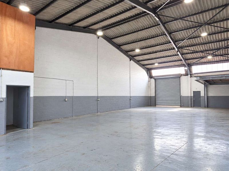 Spacious industrial warehouse unit featuring high ceilings with exposed steel trusses, a roller shutter door, mezzanine level, and durable epoxy-coated concrete flooring.