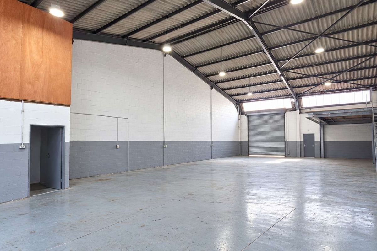 Spacious industrial warehouse unit featuring high ceilings with exposed steel trusses, a roller shutter door, mezzanine level, and durable epoxy-coated concrete flooring.