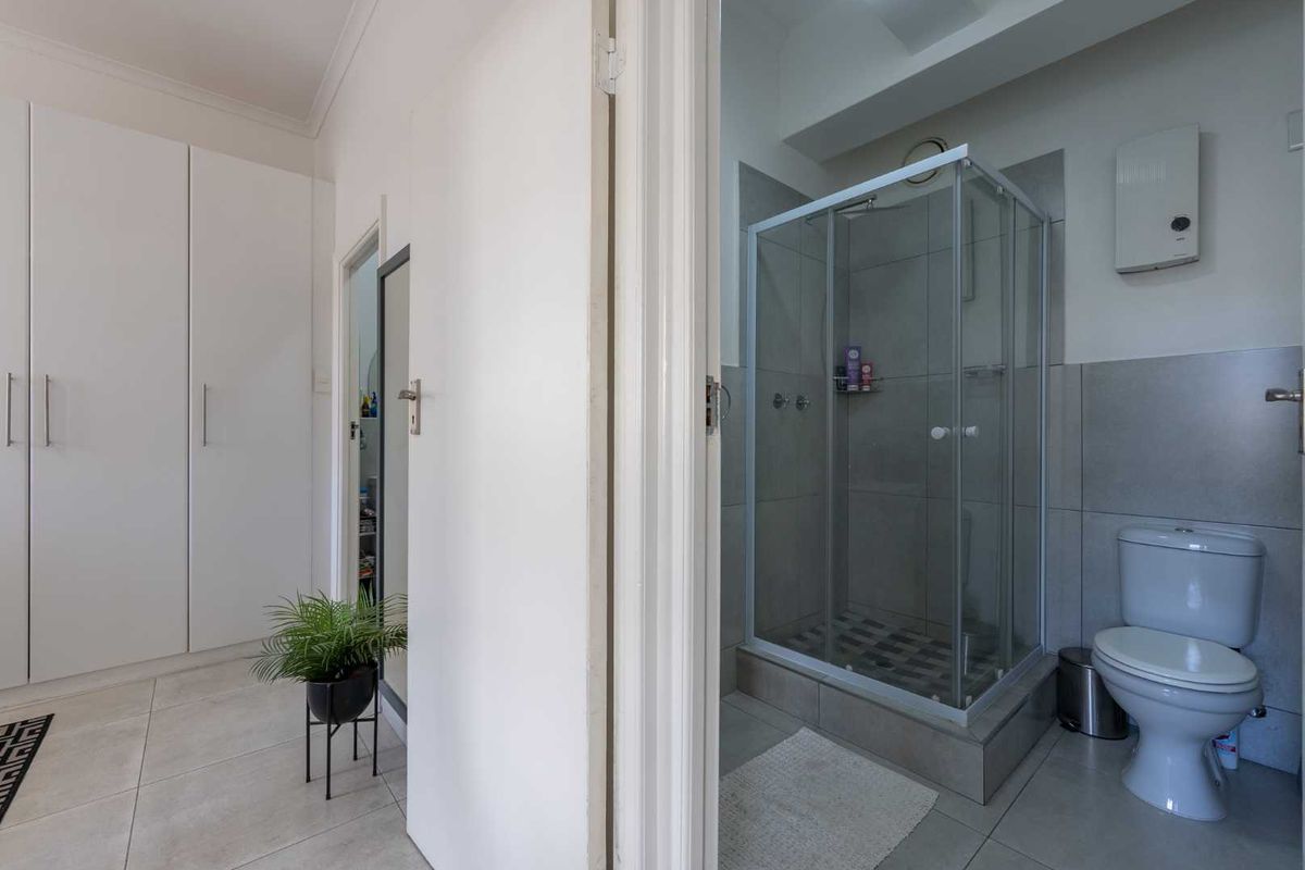 A clean bathroom featuring a glass shower enclosure, toilet, and neutral wall and floor tiling, adjacent to a hallway with built-in storage.