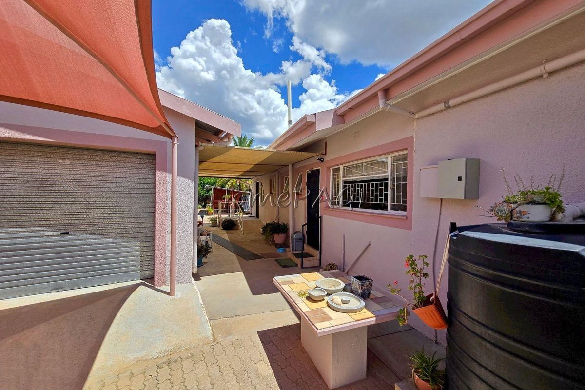 A side exterior view showing a paved walkway, a garage with a roller door, and a covered patio area with a water tank.
