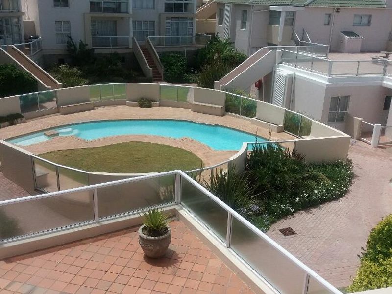 Elevated view of a communal swimming pool area featuring glass balustrades, paved patio spaces, and well-maintained landscaped gardens within the apartment complex.