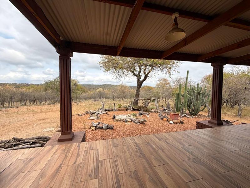 A covered patio featuring wood-look tile flooring, timber columns, and a corrugated metal roof, overlooking a landscaped garden with natural bushveld views.