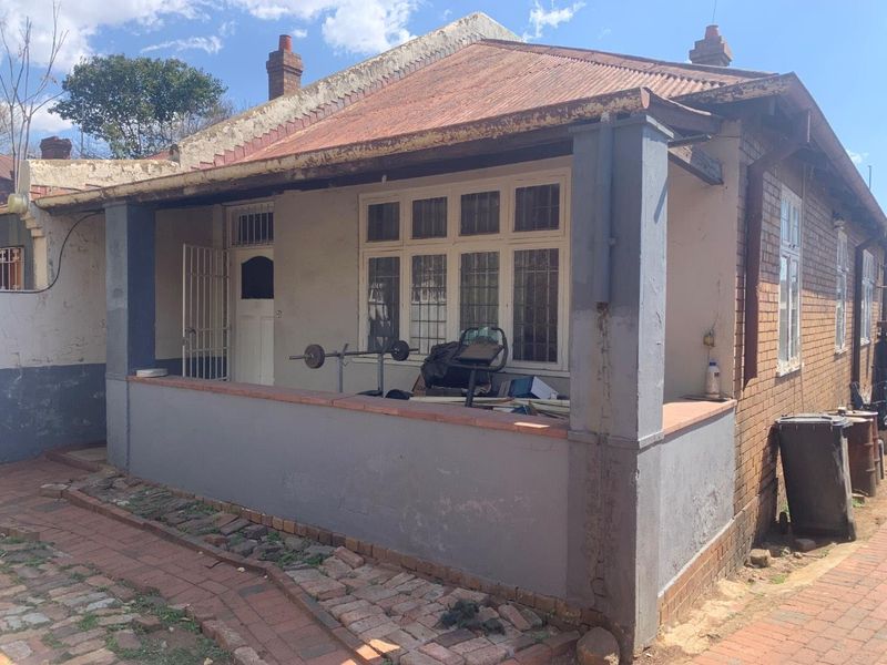 Front elevation of a traditional brick house featuring a covered porch, corrugated iron roof, and security entrance, showing signs of exterior wear.