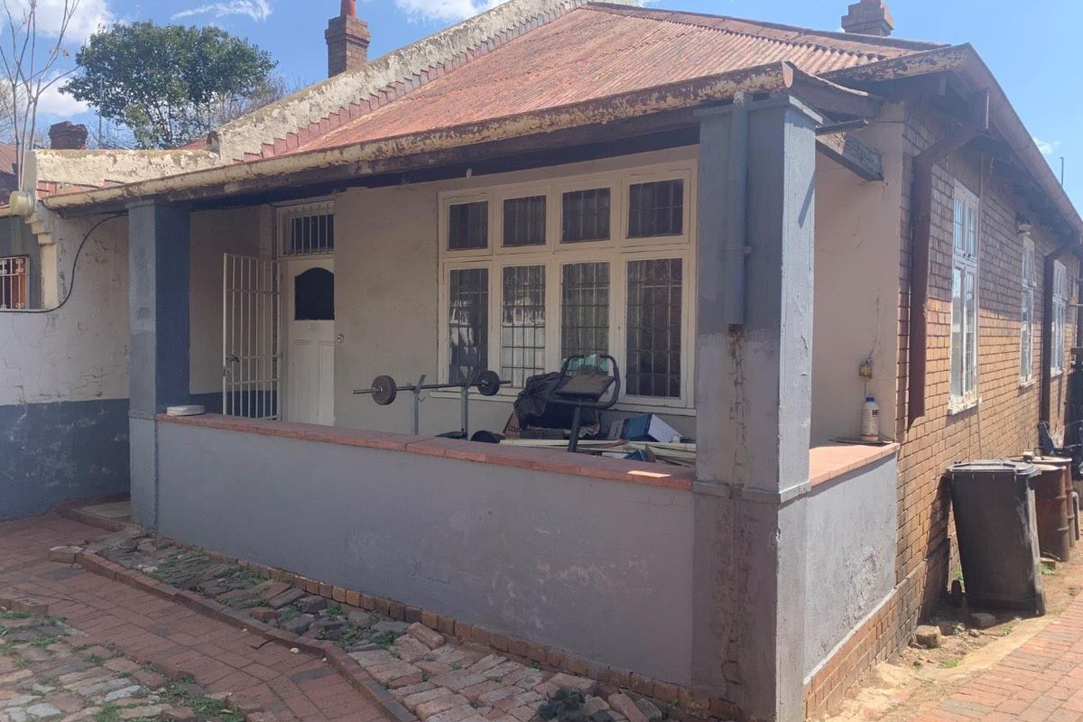 Front elevation of a traditional brick house featuring a covered porch, corrugated iron roof, and security entrance, showing signs of exterior wear.
