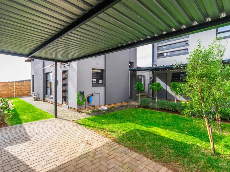 A well-maintained rear exterior featuring a paved patio area under a corrugated metal roof, a neat lawn, and secure boundary walling.
