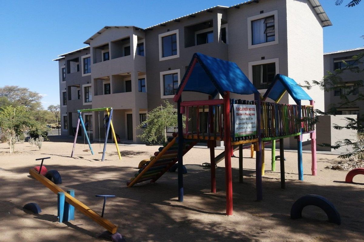 Exterior view of a multi-story apartment complex featuring a communal playground area with various play structures on a sandy surface.