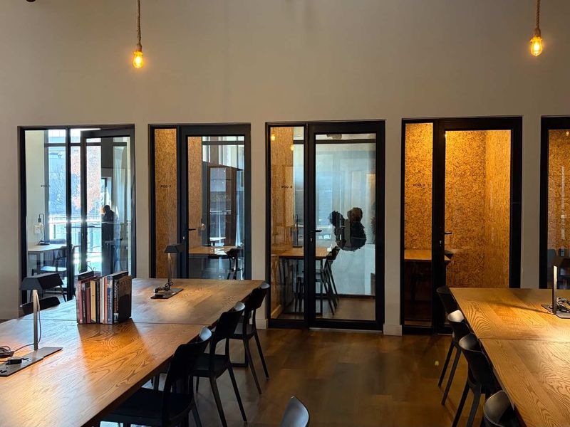 Modern office space featuring multiple glass-enclosed work pods, hardwood flooring, and integrated desk areas with contemporary pendant lighting.