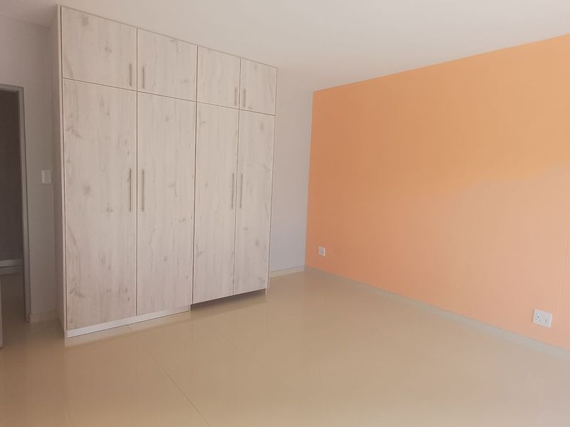 A bright, empty bedroom featuring light wood-grain built-in wardrobes, neutral tiled flooring, and a contrasting peach-toned accent wall.