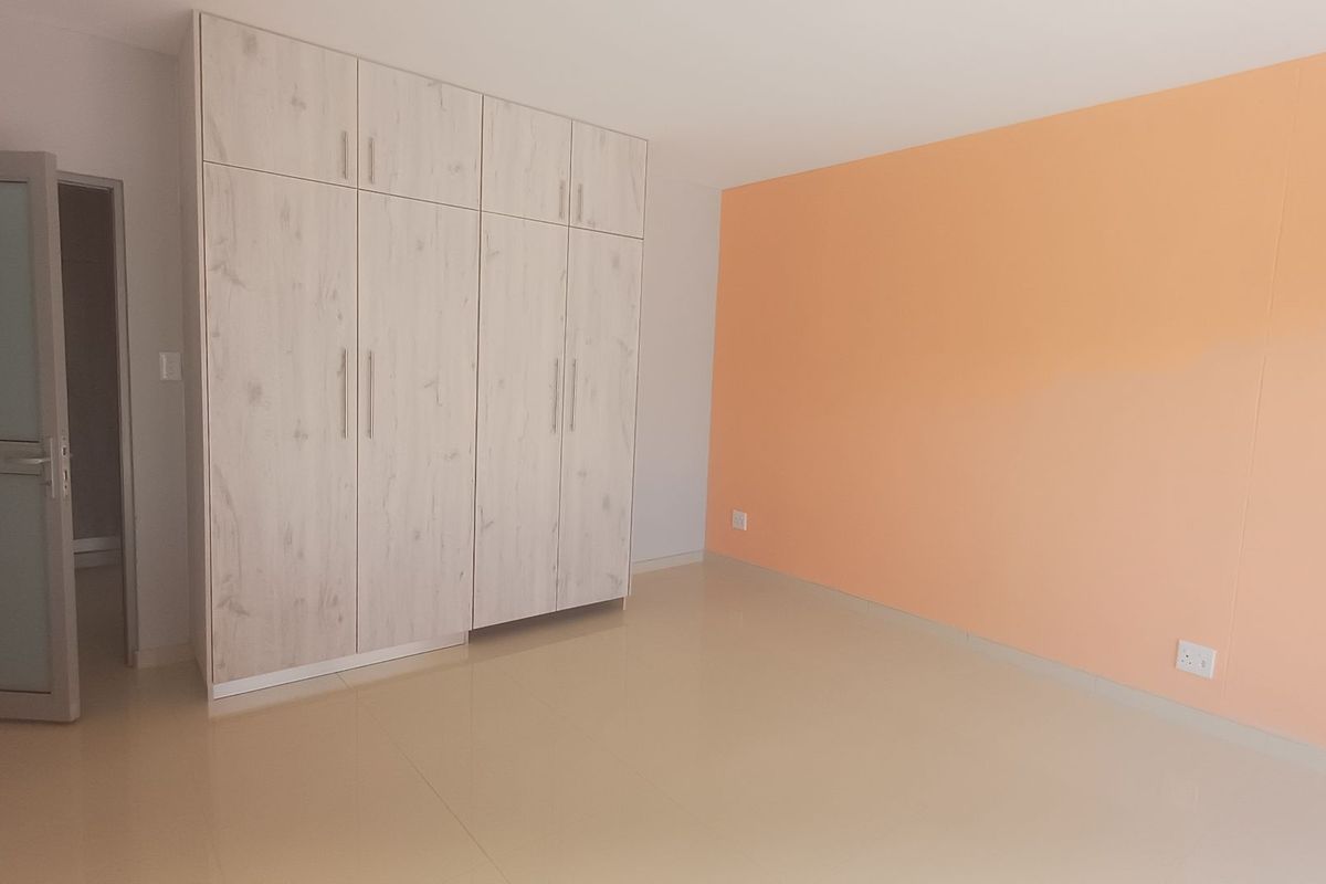 A bright, empty bedroom featuring light wood-grain built-in wardrobes, neutral tiled flooring, and a contrasting peach-toned accent wall.