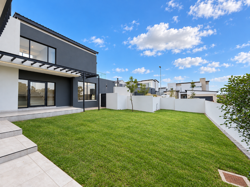 A modern rear exterior view featuring a manicured lawn, tiled patio area with a pergola, and large glass sliding doors leading into the home.