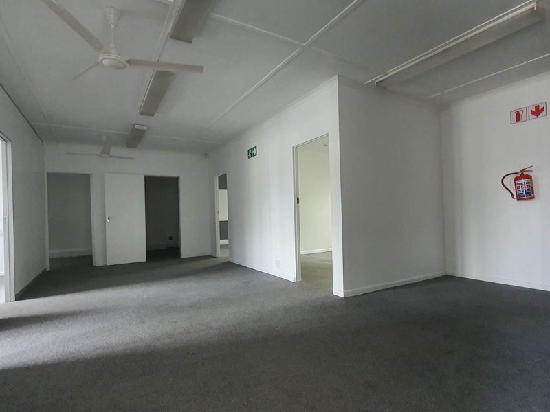 A spacious, empty office area featuring neutral walls, carpeted flooring, ceiling fans, and integrated fire safety equipment, providing a versatile workspace layout.