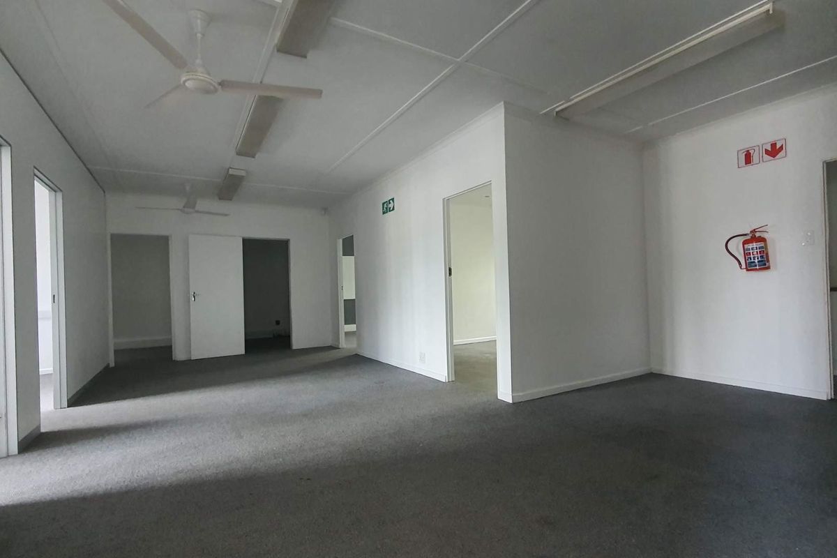 A spacious, empty office area featuring neutral walls, carpeted flooring, ceiling fans, and integrated fire safety equipment, providing a versatile workspace layout.