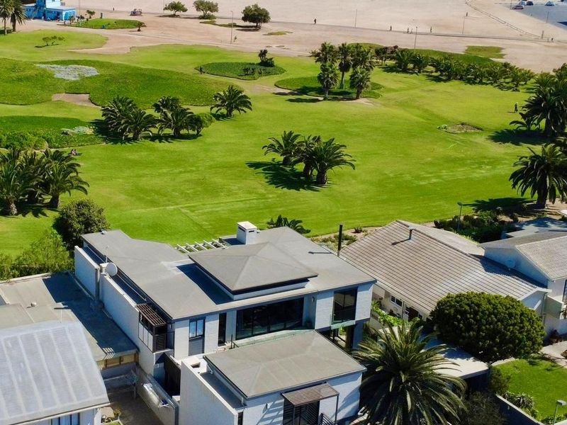 Aerial view of a modern multi-level coastal residence featuring flat roof architecture, balconies, and proximity to the beach and parkland.