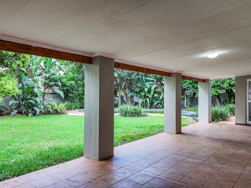 A spacious covered patio featuring tiled flooring and concrete pillars, overlooking a lush garden and swimming pool area.