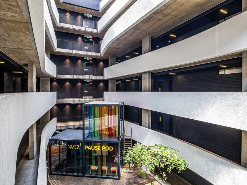 A modern multi-level apartment building atrium featuring exposed concrete architecture, metal railings, and a central glass-enclosed pause pod.