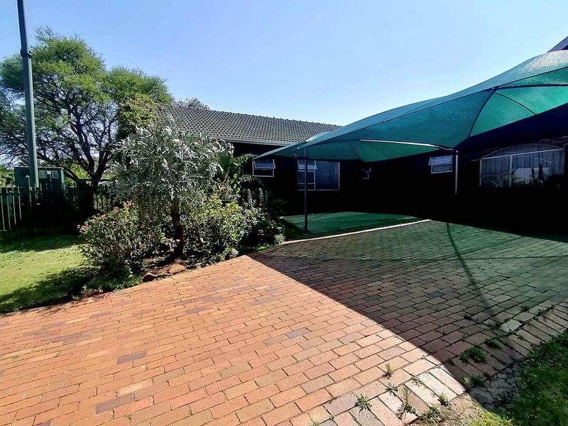 Rear exterior view featuring a brick-paved patio area covered by a green shade sail, adjacent to a garden and the main house structure.