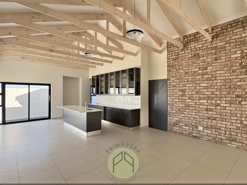 Spacious open-plan living and kitchen area featuring high vaulted ceilings with exposed timber trusses, a striking brick feature wall, and modern cabinetry.