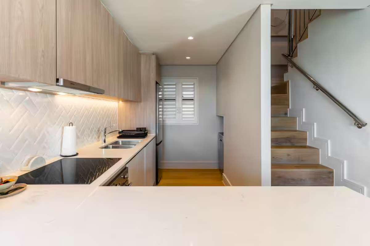 Modern kitchen featuring light timber cabinetry, stone countertops, herringbone tile backsplash, and integrated appliances, adjacent to a timber staircase with a stainless steel handrail.