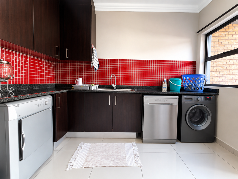 A functional laundry room featuring dark wood cabinetry, granite countertops, a striking red tiled backsplash, and tiled flooring with space for multiple appliances.
