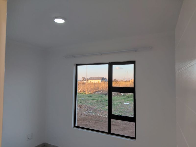 A clean, empty bedroom featuring white walls, a modern black aluminium window frame, and a recessed ceiling light, offering a bright and neutral space.