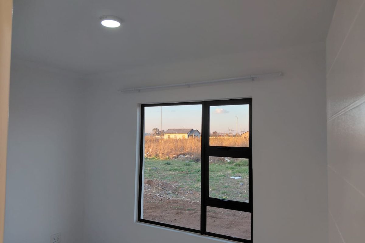 A clean, empty bedroom featuring white walls, a modern black aluminium window frame, and a recessed ceiling light, offering a bright and neutral space.