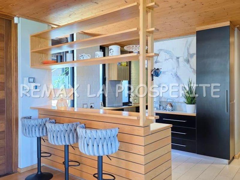 Modern kitchen featuring a timber breakfast bar with integrated shelving, sleek black cabinetry, and warm wood-paneled ceilings.