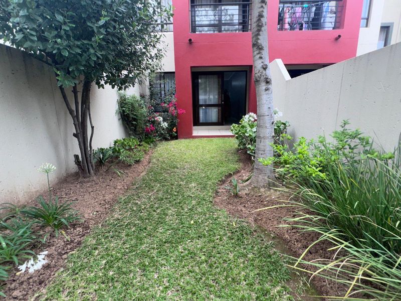 A private rear garden area featuring a small lawn, established landscaping, and access to the apartment via a sliding glass door.