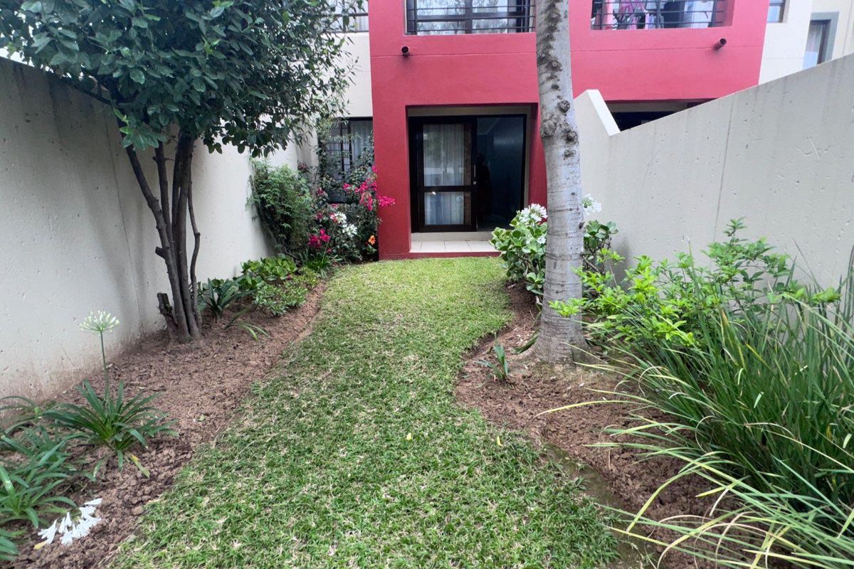 A private rear garden area featuring a small lawn, established landscaping, and access to the apartment via a sliding glass door.