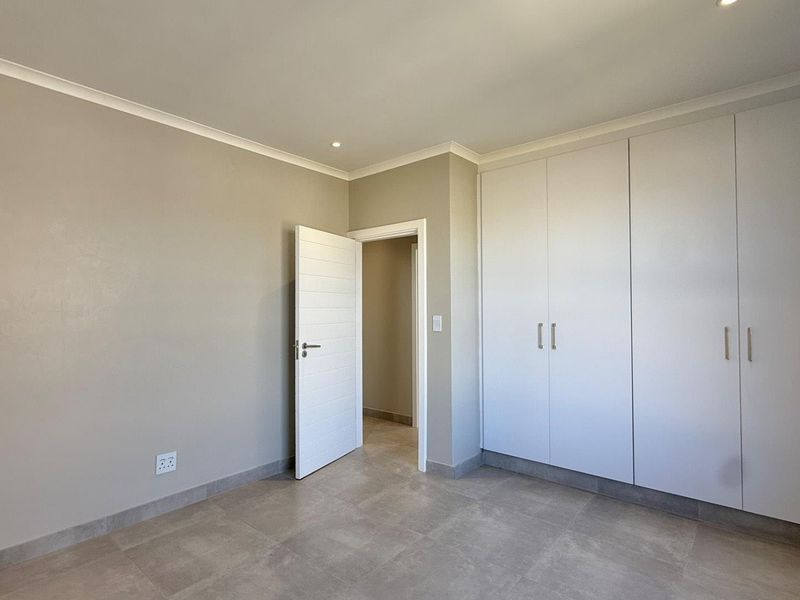 A clean, modern bedroom featuring neutral wall tones, large built-in cupboards, tiled flooring, and recessed ceiling lighting.