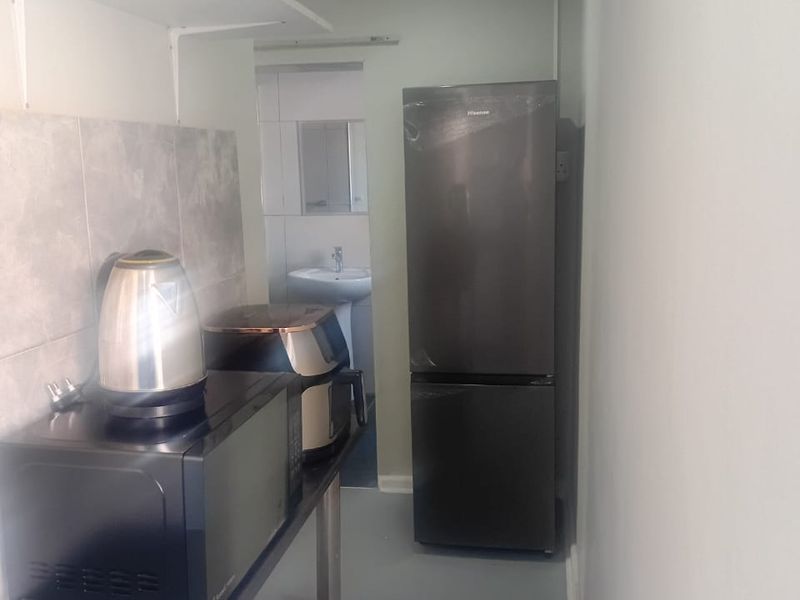 A compact kitchen area featuring a tiled backsplash, wall-mounted shelving, and direct access to an adjacent bathroom with a pedestal sink.