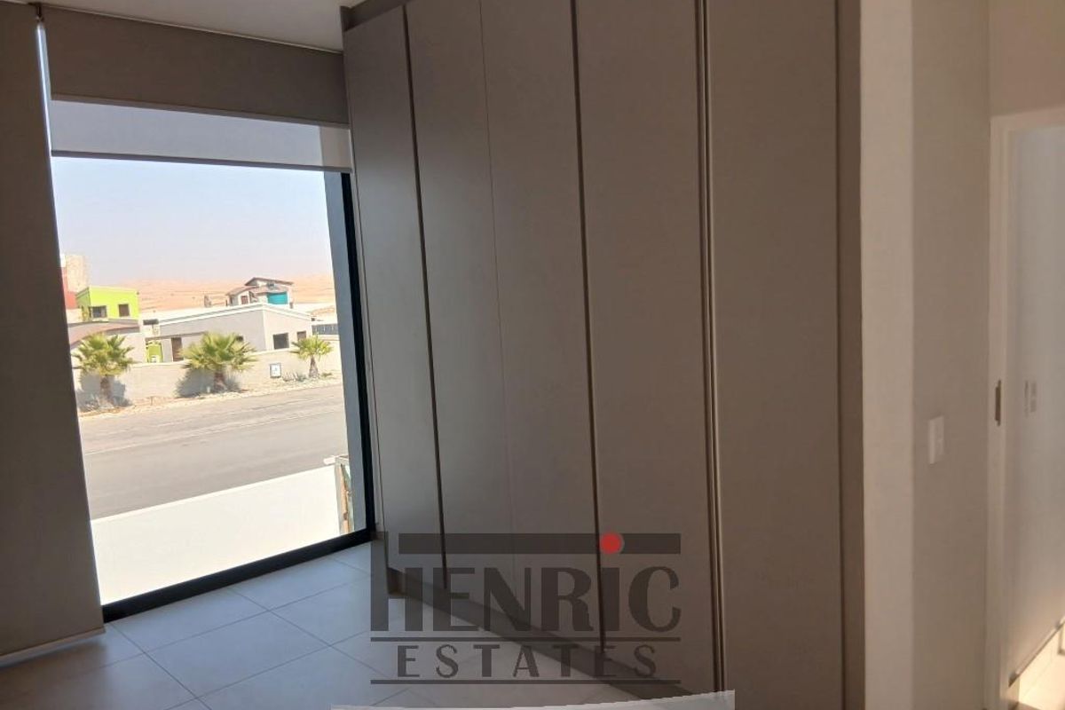 A bright, modern bedroom featuring neutral-toned built-in cupboards, large windows with roller blinds, and clean tiled flooring.