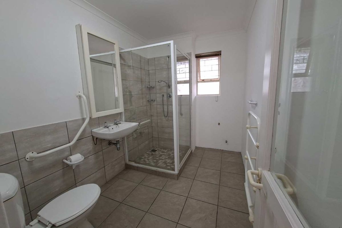A functional bathroom featuring a glass-enclosed shower, wall-mounted basin, toilet, and safety grab rails with neutral floor and wall tiling.