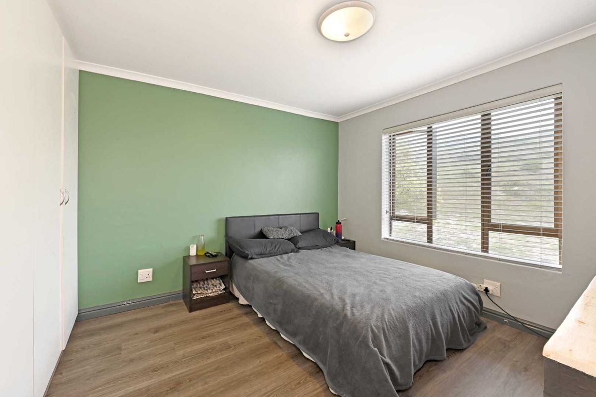 A bedroom featuring wood-look laminate flooring, a green feature wall, built-in wardrobe, and large window with fitted blinds.