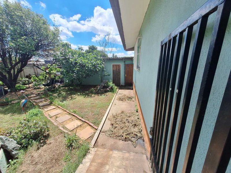 Rear exterior view showing a paved walkway, tiled patio area, security gate, and access to a small outbuilding in the garden.
